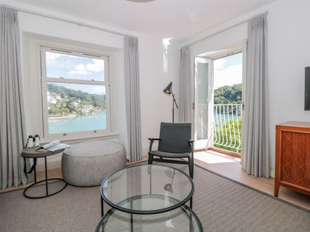 A room with a black chair a glass coffee table a round ottoman and a balcony door with a view of the water at Upper Tremorvah in Dartmouth