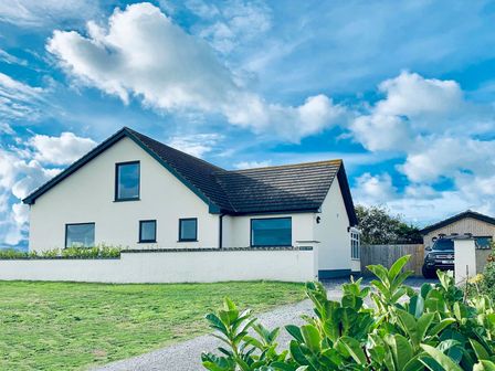 A white house with a dark roof behind a low wall with bushes in front and a car parked near a wooden garage at Hafod Heli in Rhosneigr