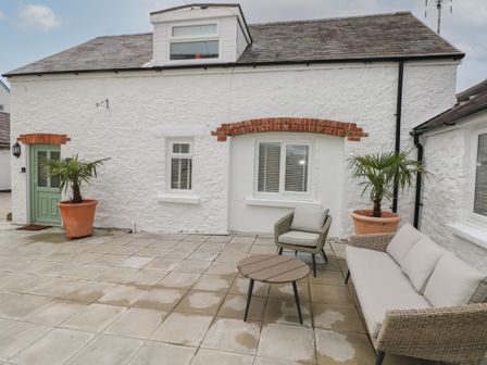 An outdoor patio with beige cushioned wicker sofa and chair a round wooden table and potted palm plants in front of a white stone house with green door at Rosemary Cottage Jameston near Manorbier