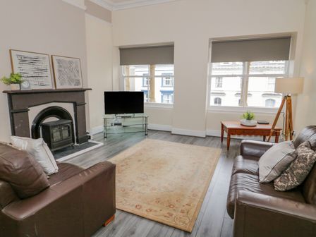 A living room with two brown sofas a fireplace a TV on a glass stand a wooden table and two windows with gray blinds at Gerrard House Apartment in Llandudno