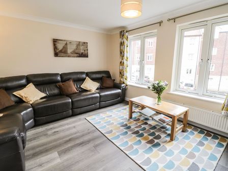 A living room with a black leather sofa, wooden coffee table with flowers, patterned rug, and windows at Keel House in Whitby