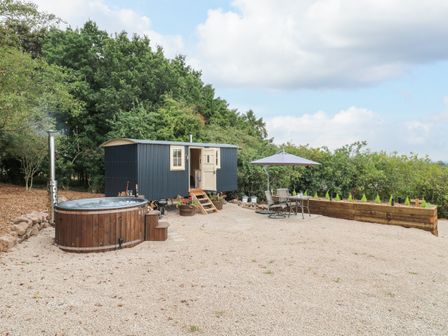 An outdoor area with a wooden hot tub a small black shepherds hut with steps a table with chairs and an umbrella at High Grounds Shepherd's Hut Boylestone near Ashbourne
