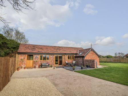 A single story brick building with wooden doors and windows a patio with chairs and potted plants and a grassy lawn at The Old Piggery in Defford
