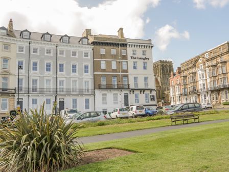 A row of buildings with parked cars in front and a grassy area with a bench at Foxton Penthouse in Whitby