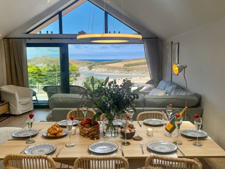 A dining table set with plates glasses and food in a room with large windows and a view of hills and a beach at Porth Beach in Porth near St Columb Minor