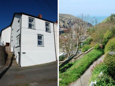 A white two-story house with stairs outside and a stone path with greenery and a view of a hill and sea at Glan y Mor in Porthgain near St Davids