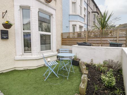 A small outdoor patio with a blue table and two chairs on artificial grass next to a flower bed at Apartment 2 - Llandudno Central in Llandudno