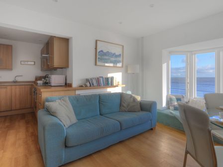 A living room with a blue sofa and pillows next to a kitchen with wooden cabinets and a window with a sea view at Sea Fever in Lyme Regis