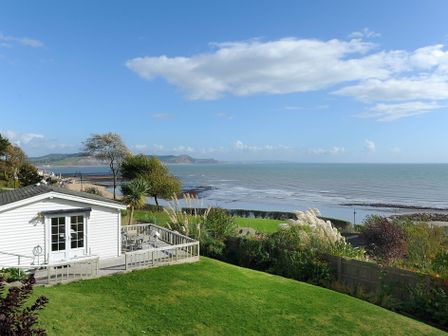 A white cabin with a wooden deck and furniture overlooking the sea with grassy lawn and trees at The Cabin in Lyme Regis