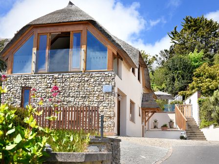 A house with stone and plaster exterior and a thatched roof surrounded by plants and trees at Bucket & Spade in Lyme Regis