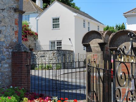 A street with a white house behind a stone wall and iron gates with flowers in the foreground at Blacksmiths Cottage Lyme Regis