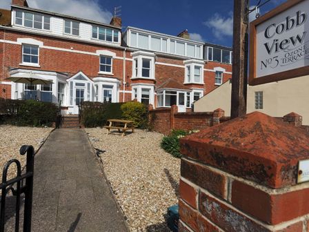 A brick house with white window frames and a paved walkway leading to the front door at 1 Cobb View Lyme Regis