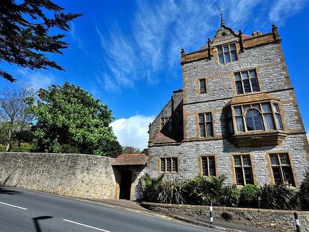 A stone building with multiple windows and a bay window near a road with a stone wall and trees at 6 Coram Tower in Lyme Regis