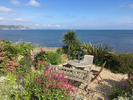 A garden with a wooden table and chairs overlooking the sea with flowers and greenery at 4 East Cliff in Lyme Regis