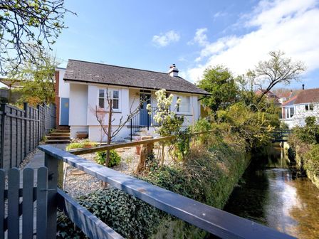 A small white house with a grey roof next to a narrow river and a wooden fence at The Bolthole in Lyme Regis