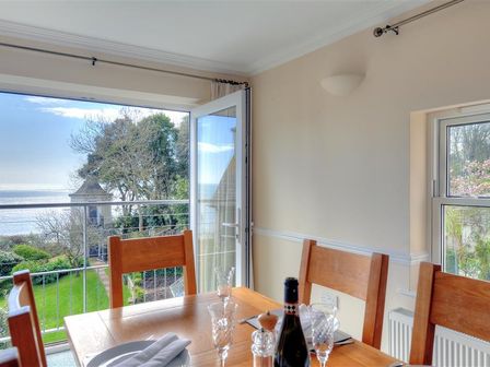 A dining room with wooden chairs a table set with glasses and a bottle with view of garden and sea through open door at The Portland Suite in Lyme Regis