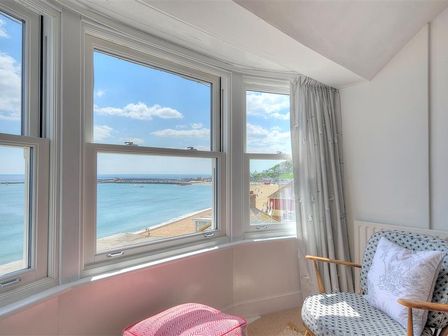 A corner room with a patterned armchair a pink footstool and large windows overlooking the sea at Ross House in Lyme Regis