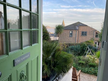 A green door with a sign saying The Apartment opening to a garden with shrubs a wooden chair and a brick building in the background at The Apartment at Queen Anne's Lodge in Lyme Regis