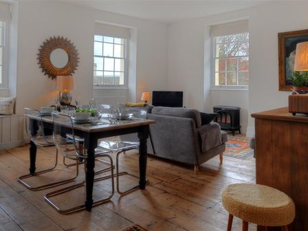 A dining and living area with a wooden table, clear chairs, gray sofas, and windows at 2 Woodville Lyme Regis