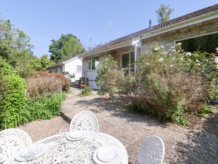 An outdoor patio area with a white metal table and chairs set with plates and glasses next to a stone house with flowering bushes at High Trees in Lyme Regis