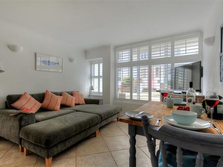 A living room with a gray sectional sofa and striped pillows next to a wooden dining table set for two with wine glasses and plates at Waterloo Place in Charmouth