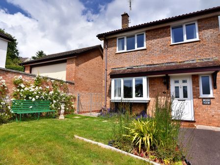 A brick house with a white door and windows a front garden with a green bench a flower bed and a statue at Woody View in Colyton
