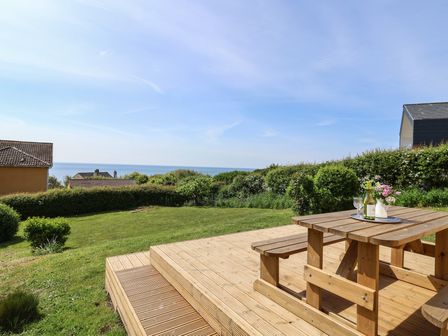 A wooden picnic table on a deck overlooking a grassy garden with bushes and houses near the sea at Tillicum in Charmouth