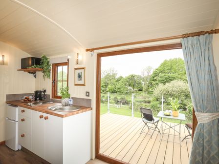 A small kitchen with a sink and countertop next to a glass door leading to a wooden deck with a table and chairs overlooking trees at Llety'r Bugail 2 in Llanrug