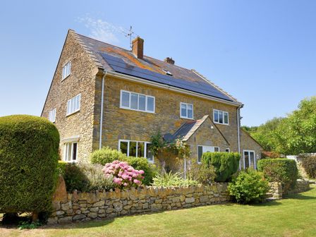 A stone house with solar panels on the roof surrounded by a garden and bushes at Manor Lodge Burton Bradstock