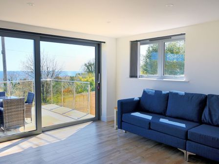 A living room with a blue sofa wooden floor and large glass sliding doors overlooking a balcony and trees at Yarrow in Charmouth