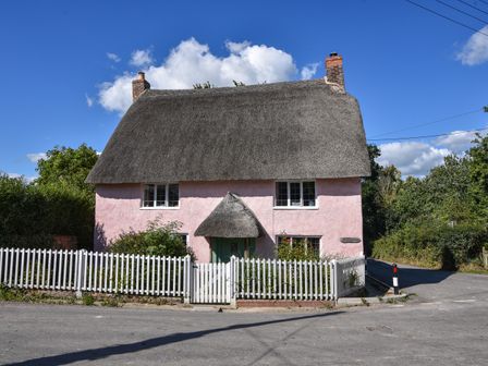 A pink thatched roof cottage with white picket fence and greenery at Old Cross Cottage in Charmouth