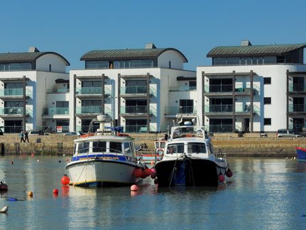 Two boats moored in a harbor with modern apartment buildings and parked cars in the background at Harbour View in West Bay