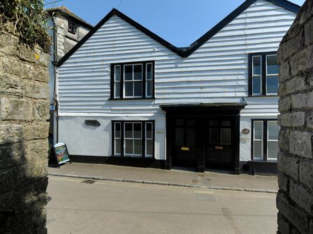 Exterior view of a white timber-clad building with black framed windows and doors on a street with stone walls on either side Captains House Lyme Regis