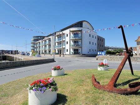 A street corner with a large rusted anchor and flower pots in front of modern apartment buildings at Sandstones in West Bay