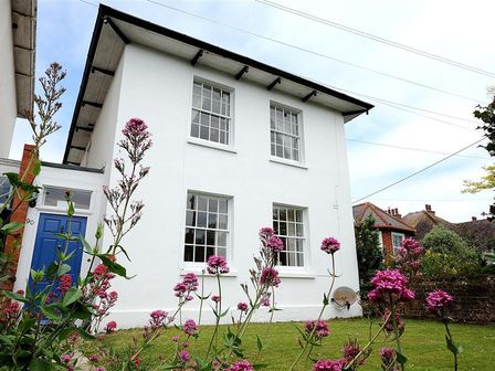 A white two-story house with four windows and a blue door surrounded by green grass and pink flowers