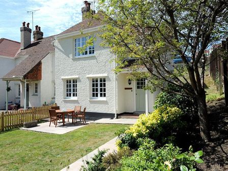 A white cottage with a garden table and chairs on a patio and green plants and trees in the yard at 1 Riverside Cottages Lyme Regis