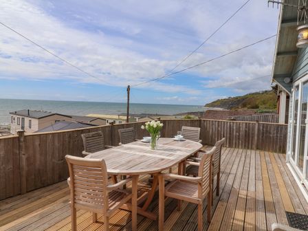 An outdoor wooden deck with a wooden dining table and six chairs with a vase of flowers on the table overlooking the sea at Ammonite in Lyme Regis