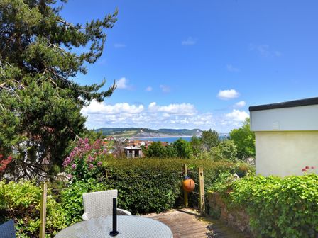 A garden patio with a round glass table and chairs overlooking trees and hills with a view of the sea at Little Bay View in Lyme Regis
