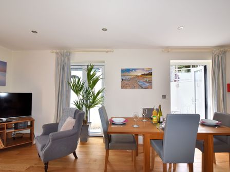 A dining area with a wooden table set with plates and glasses and a gray armchair near a tv at Seas The Day in Lyme Regis