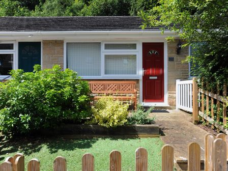 A front garden with bushes and a wooden bench in front of a house with a red door at 36 Fernhill Heights in Charmouth