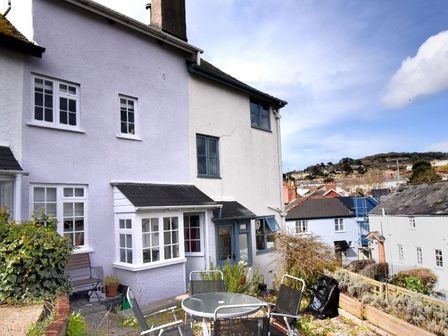 An outdoor patio area with a glass table and chairs outside a house with white and light purple walls at Lavender Cottage in Lyme Regis