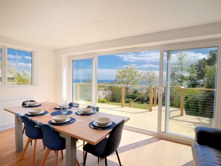 A dining area with a wooden table set with plates and glasses next to large sliding glass doors opening to a deck with trees and sea view at Lottys Lookout in Lyme Regis