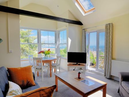 A living room with a wooden coffee table, sofa with cushions, dining table with chairs and flowers, TV, large windows and glass doors at Bay View in Lyme Regis