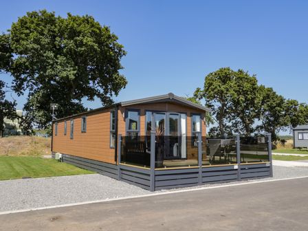 A wooden lodge with glass doors and a fenced patio on a gravel driveway with large trees nearby at Away Daze in Carnaby near Bridlington