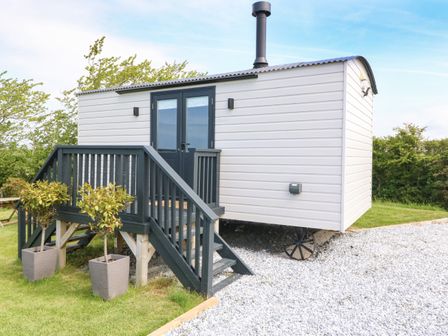 A small white cabin with black double doors and a chimney on a gravel area with stairs and plants at Cwt Y Rhos in Valley