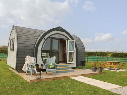 A pod cabin with open door and two chairs on a wooden porch with picnic table and green fence in background at Poppy in Seaton near Hornsea