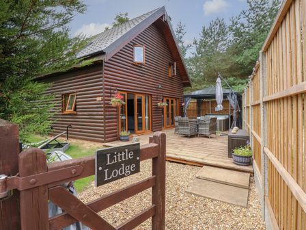 A wooden lodge with a deck patio and outdoor furniture behind a gate with a little lodge sign in Pentney near Kings Lynn