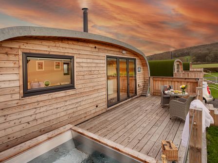 A wooden cabin with a deck patio area featuring a hot tub a round table with chairs and a scenic view at Middle Longlands in Cartmel