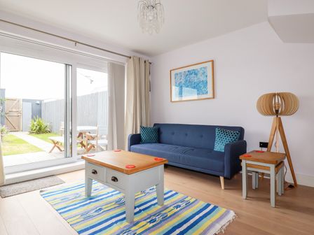A living room with a blue sofa two blue cushions a wooden coffee table a floor lamp and glass sliding doors leading to a garden at 6 Ocean Heights in Newquay