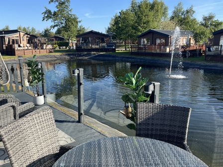 An outdoor area with a table, chairs, and a view of cabins near water at Loluke Lodge on The Lake, 5 Coniston Drive, Borwick near Carnforth